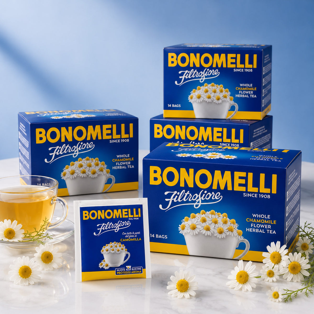 Four blue and yellow boxes of Bonomelli Filtrofiore with whole chamomile flower herbal tea arranged on a blue background, with a single tea bag packet standing in front and small white chamomile flowers scattered around.