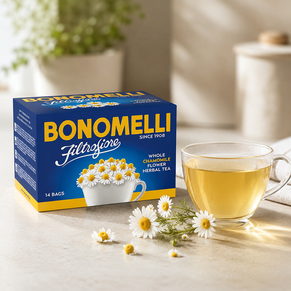 A box of Bonomelli Filtrofiore with whole chamomile flower herbal tea sits on a light stone surface next to a clear glass cup filled with golden tea, with a few chamomile flowers scattered in front. The background is softly blurred with warm sunlight and neutral decor.