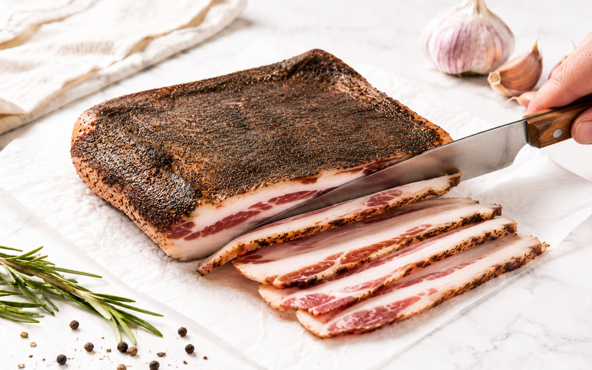 Guanciale being sliced on white parchment paper with visible marbled meat and fat, black pepper crust, rosemary, garlic, and marble background