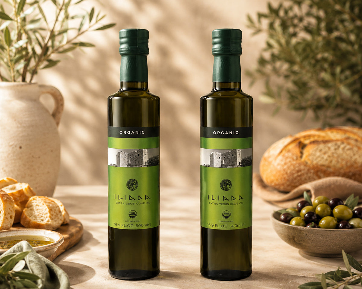 Two bottles of Iliada Organic Extra Virgin Olive Oil standing on a wooden surface, surrounded by olive branches, bread, and olives in a warm, rustic setting.