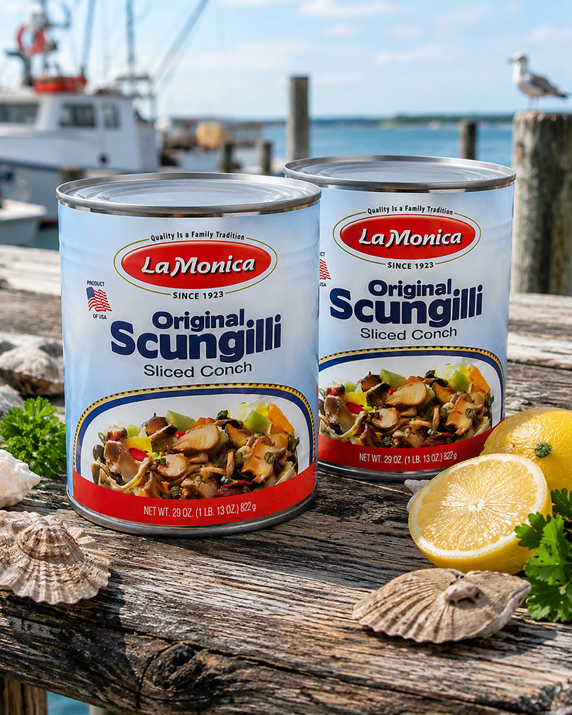 Two cans of LaMonica Original Scungilli Sliced Conch placed on a rustic wooden dock with seashells, lemon halves, parsley, and a blurred marina background with boats and water.