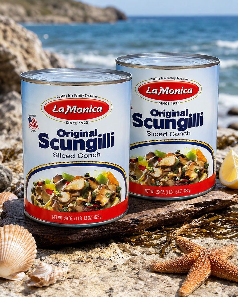 Two cans of LaMonica Original Scungilli Sliced Conch placed on a rocky beach with starfish, shells, seaweed, and ocean water in the background.