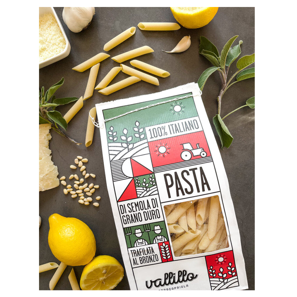 Vallillo Penne Lisce pasta package with uncooked penne, lemon, garlic, and herbs on a countertop.