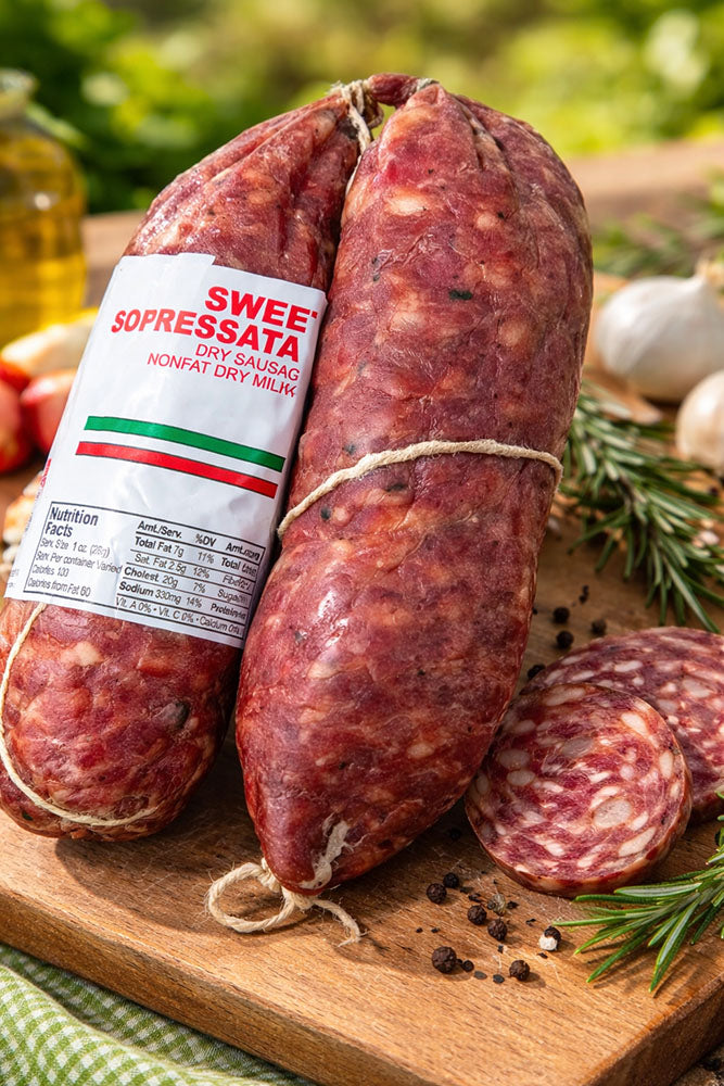 Two Alps SWEET SOPRESSATA sausages on a wooden board with herbs and garlic, featuring a visible label.
