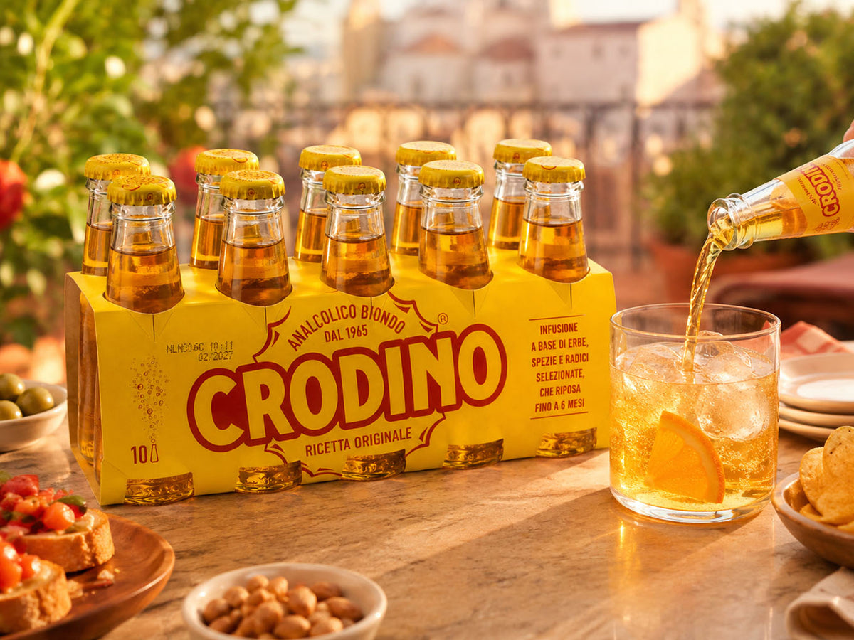 A 10-pack of Crodino non-alcoholic aperitif glass bottles on a table, with a bottle pouring crodino into a glass with an orange slice in an outdoor dining setting.
