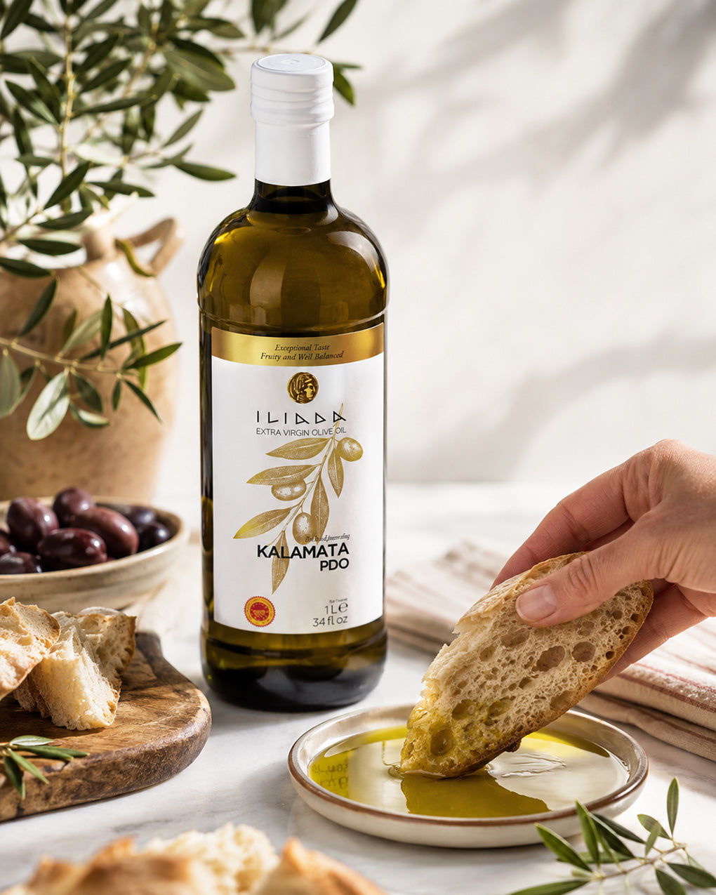 A bottle of Libbk Kalamata 100% olive oil stands on a light marble surface beside a hand dipping a slice of bread into a small dish of olive oil. Olives, bread, and olive branches are arranged around it in a bright, styled kitchen scene.
