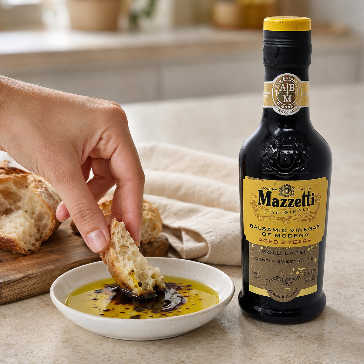 A bottle of Mazzetti Originals Balsamic Vinegar of Modena stands next to a hand dipping a piece of bread into a small white bowl containing the dark balsamic, and olive oil with rustic bread.