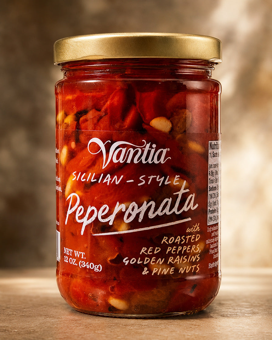 A jar of Vantia Sicilian-Style Peperonata, featuring roasted red peppers, golden raisins, and pine nuts in a clear glass jar with a gold lid and white label text, set against a warm, blurred background.