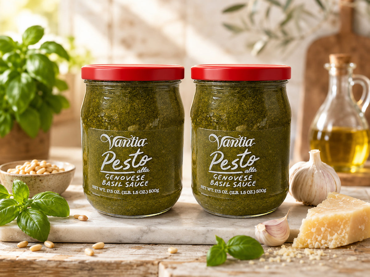 Two jars of Vantia Pesto alla Genovese Basil Sauce with red lids, displayed on a rustic wooden table with fresh basil, garlic, parmesan cheese, and a bottle of olive oil.